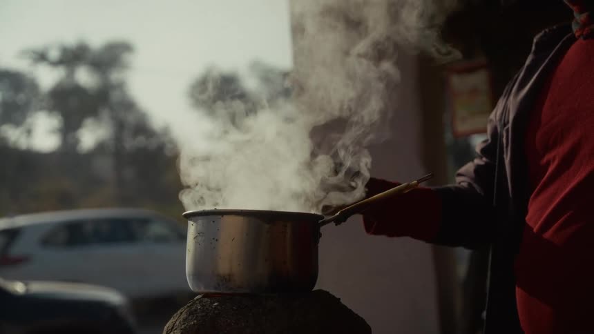 A black man wearing dark clothing is standing over a small silver pot of liquid vigorously boiling on a makeshift burner with steam billowing into the air and the blurred background showing a busy outdoor street with vehicles