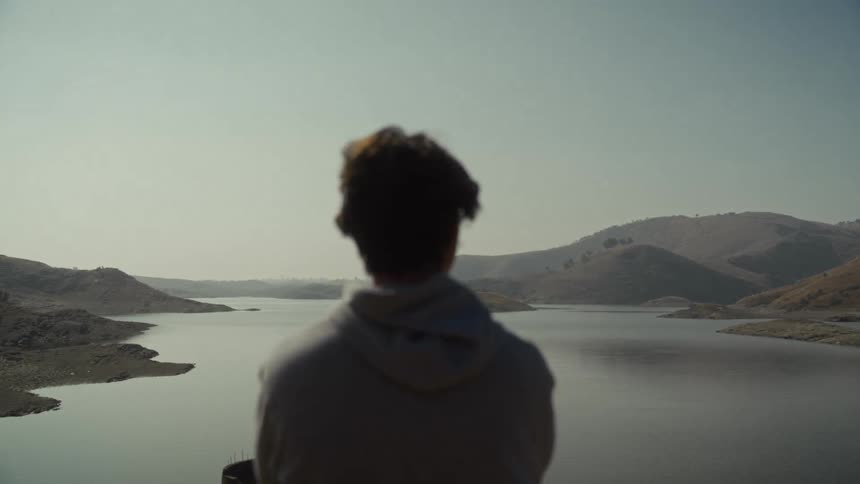 A person in a lightcolored hooded sweatshirt is seen from behind slightly out of focus looking out over a wide still body of water surrounded by dry rolling hills under a bright hazy sky