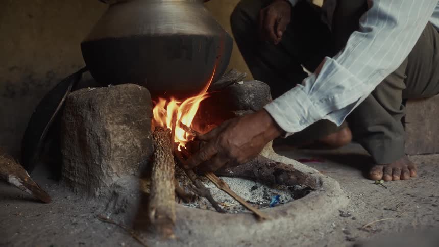 A man wearing a lightcolored longsleeve shirt and dark pants squats beside a traditional stone fire stove and feeds small pieces of wood into the burning fire below a large blackened cooking pot