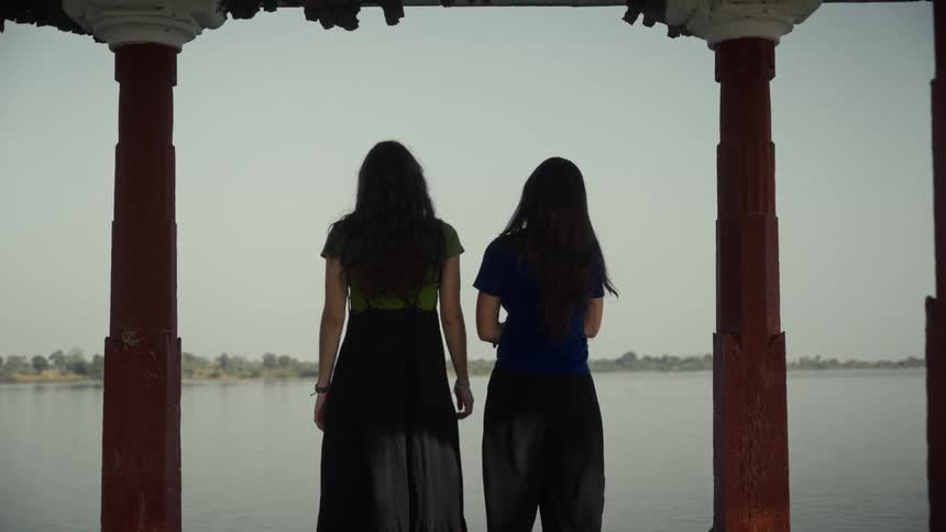 Two women with long dark hair are standing under a redpillared structure looking out at a calm lake or river that fills the backgroundthe women are wearing casual dark clothingone in a green and black outfit the other in a blue top and dark pants