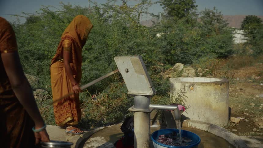 A woman in a vibrant sari operates a manual hand pump as another waits patiently in the foreground with a metal pot to collect water in a sundrenched village