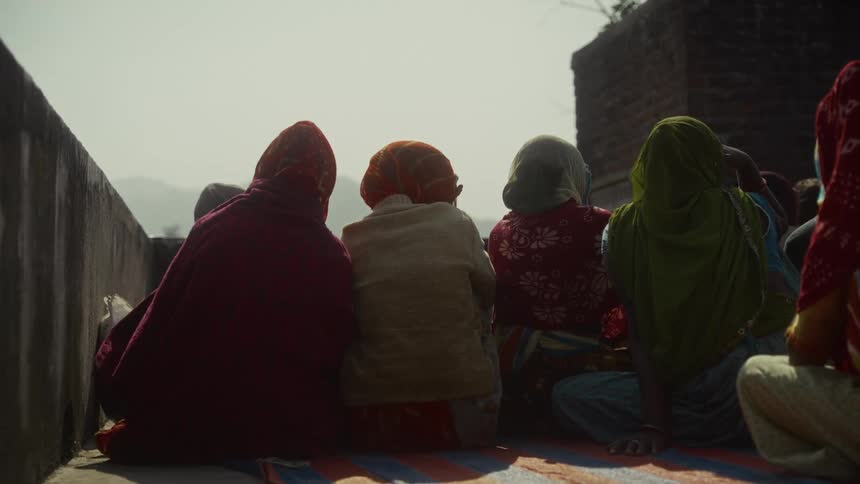A tight shot focuses on the backs of several women who are seated closely together on a striped mat and wearing traditional head coverings in various colors as they face the hazy horizon from a brightly sunlit rooftop