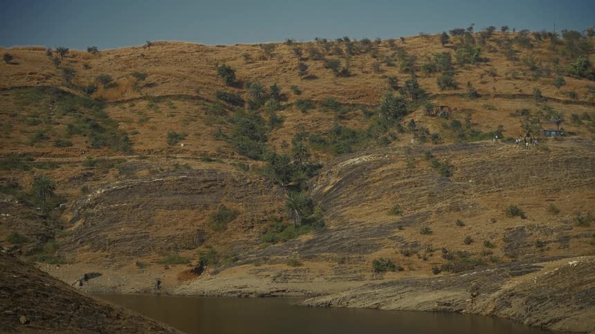 A wide landscape view features a dry tiered brown hillside covered in sparse scrub and scattered small trees sloping down to meet a body of dark water in the foreground all under a clear blue sky