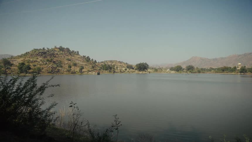 A lake lies in the countryside at midday surrounded by rolling hills and expansive open land