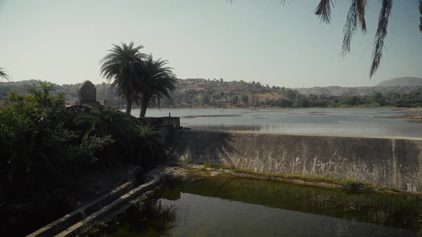 Reservoir bordered by vegetation and concrete structure with dry hills in the background under clear sky