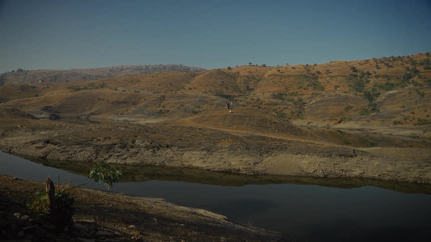 A wide static shot captures a large calm body of dark water surrounded by extensive brown dry and sparsely vegetated hillsides under a clear blue sky