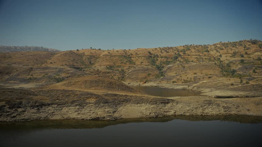 A vast dry landscape features terraced brown hills covered in sparse vegetation sloping down to meet the edge of a small dark body of water under a clear blue sky