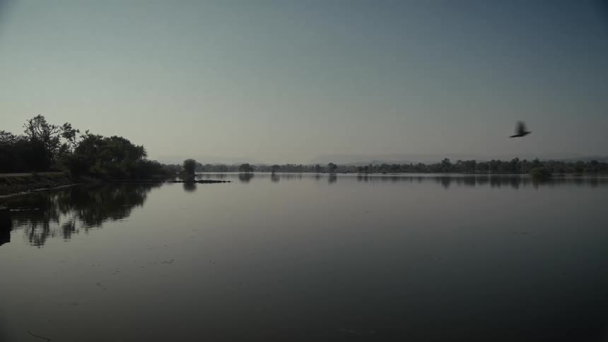 A wide tranquil body of water reflects the faint trees along its distant bank and the pale hazy sky above creating a symmetrical muted landscape
