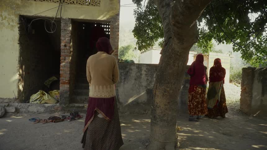 Some women wearing traditional long skirts and head coverings stand and interact in the dusty yard of a simple aging building with a large textured tree trunk partially obscuring the view