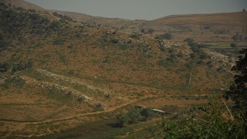 The video captures a sweeping view of dry rocky hillsides covered in sparse brown and green vegetation the landscape features rugged terrain and appears to be in a rural possibly arid or semiarid region