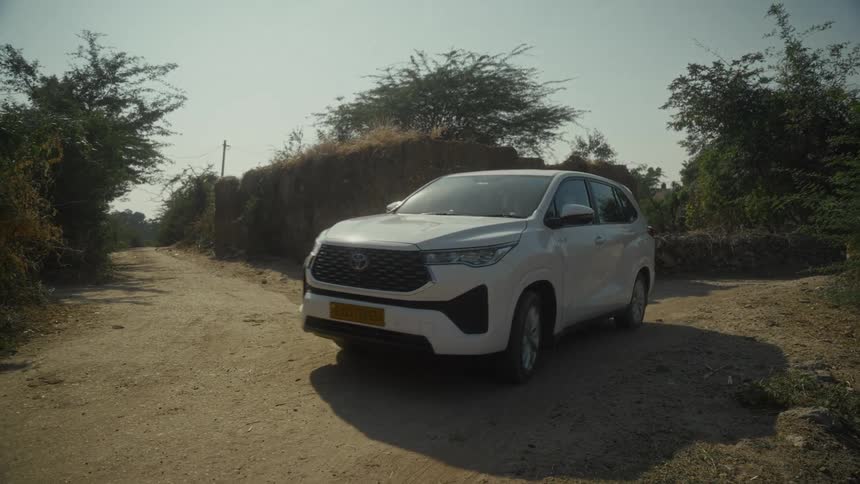 A white suv is driving slightly off a fork in a dry unpaved road next to a large rough mound of earth with dry scrub brush and trees growing around the edges under a bright hazy sky