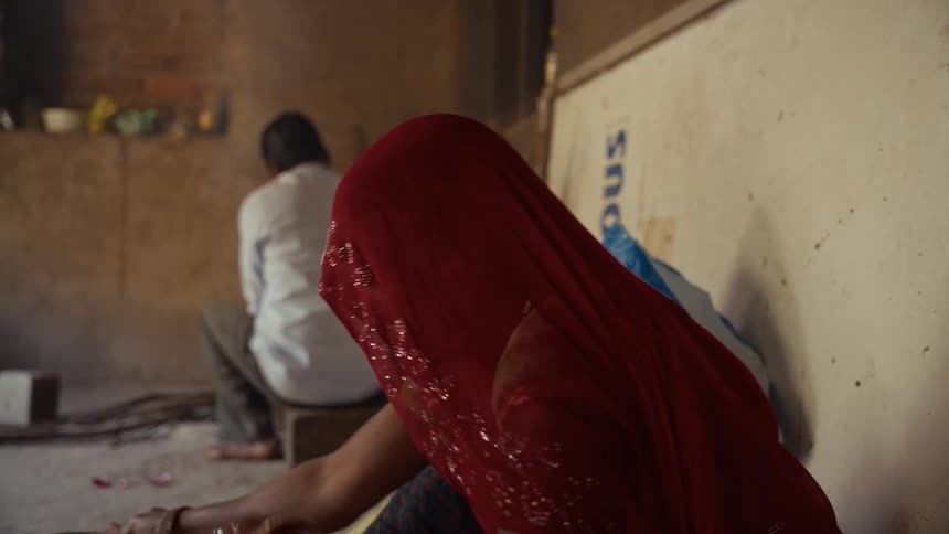Person with red veil holding metal container in modest setting with another person in the background