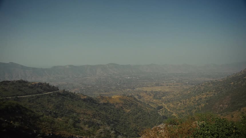 A wide static shot captures a sprawling mountain valley view showing layers of rugged vegetated hillsides sloping down to a distant valley floor all under a vast pale blue sky