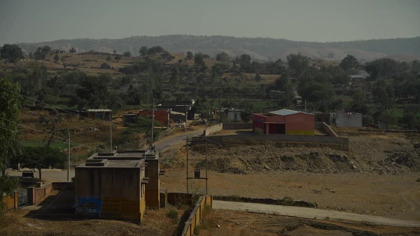 An elevated view of a small rural town with scattered buildings nestled in a dry dusty valley surrounded by tree covered hills under a hazy sky