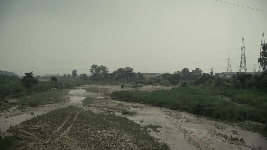 The video displays a wide dry riverbed with shallow muddy water flowing through multiple small channels the landscape is flat and hazy with power line towers visible on the right side and a line of trees in the distance
