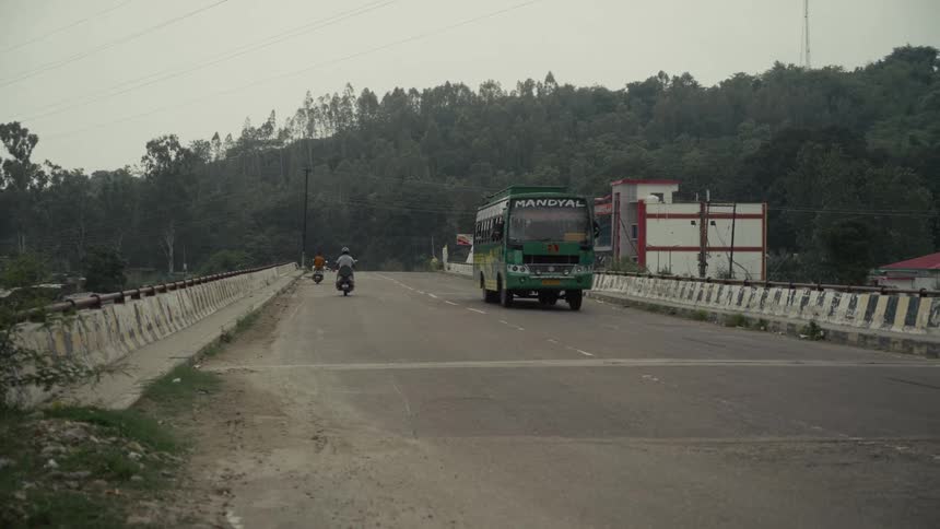 A green bus drives past the camera on a bridge road passing motorbikes heading toward a forested hillside