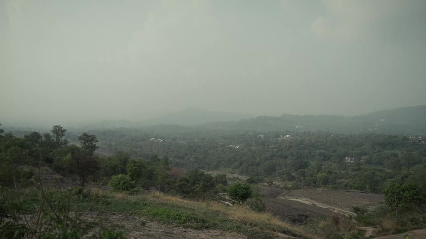 A hillside in the countryside stretches into the distance with vast land covered in green vegetation and a few scattered displaced buildings dotting the landscape