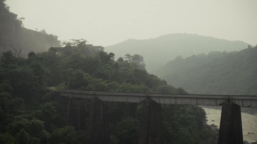 The video shows a long elevated concrete bridge or viaduct spanning a steep heavily forested ravine and a river below the scene is misty and overcast with layers of green hills receding into the distance