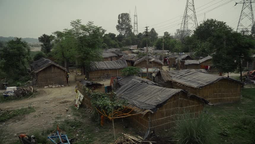 A cluster of temporary shelters or huts constructed from materials like straw and thatch with dark coverings for roofs is nestled in a dusty open area surrounded by treesin the background tall metal electricity pylons and power lines loom over the