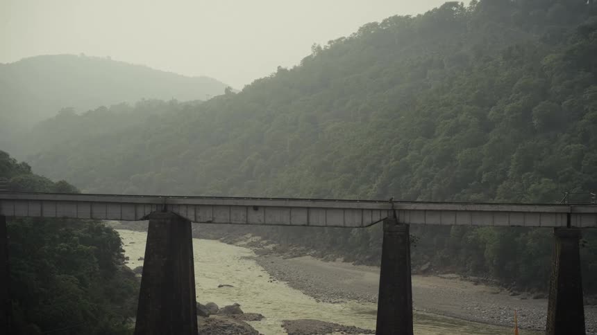 Concrete bridge over river in mountainous area with dense vegetation and mistcovered hills in the background