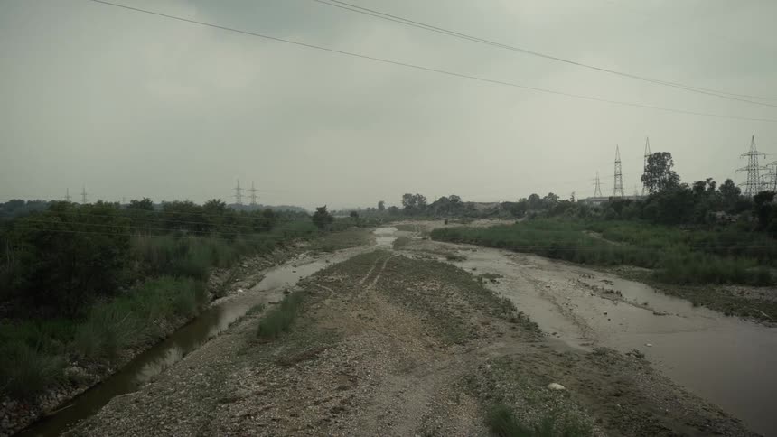 Muddy streams of water over soil covered in gravel surrounded by bushes with buildings and transmission towers in around outdoors during day time
