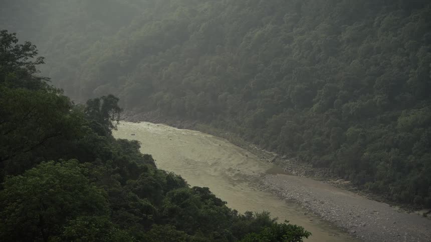 A wide muddy riverbed and fastmoving brown water snaking through a steep valley densely covered in lush dark green forest