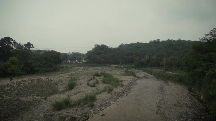 The video is a static wide shot of a dry riverbed or a stream with very low water levels composed mostly of gravel and small rocks the banks are covered in dense dark green foliage leading up to a horizon dominated by forested hills under a hazy sky
