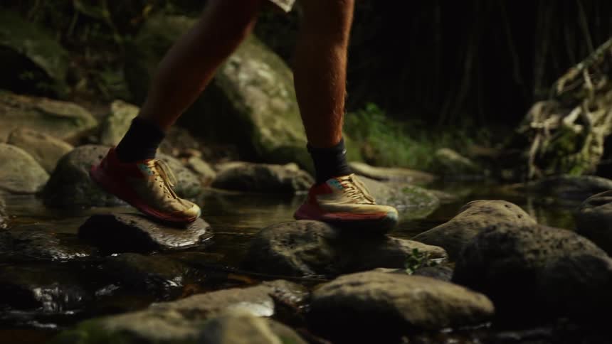 A person wearing colorful running shoes steps carefully across wet rocks in a stream