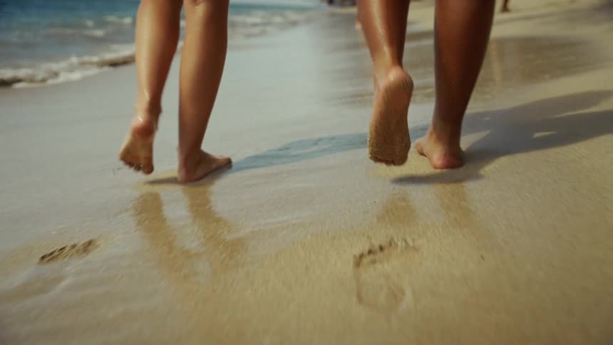 Female lower legs and feet of two people walking on wet sand next to the sea at the beach towards other people on the background outdoors during day time
