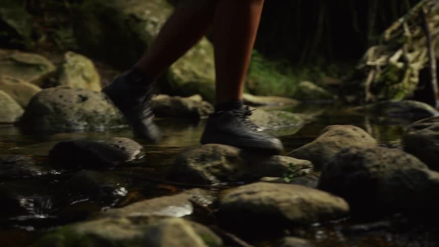 Person wearing hiking shoes stepping on stream rocks surrounded by vegetation