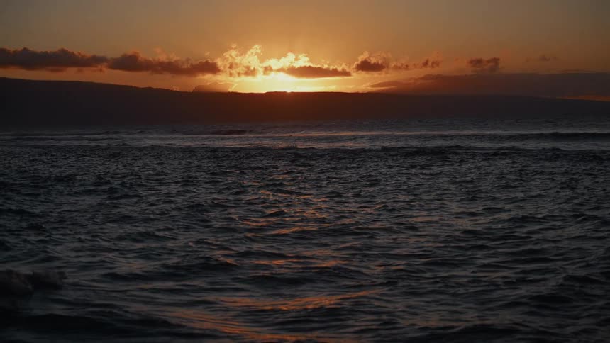 Sun sets behind clouds and distant mountains tinting sky and sea with golden hues