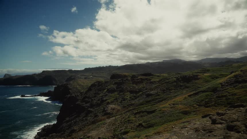 A sweeping view pans across a rugged green landscape revealing a dramatic coastline where the deep blue ocean crashes against rocky cliffs under a vast cloudy sky
