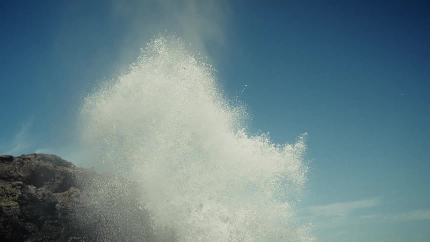 A rocky coastline at midday features ocean water forcefully springing up from a blowhole