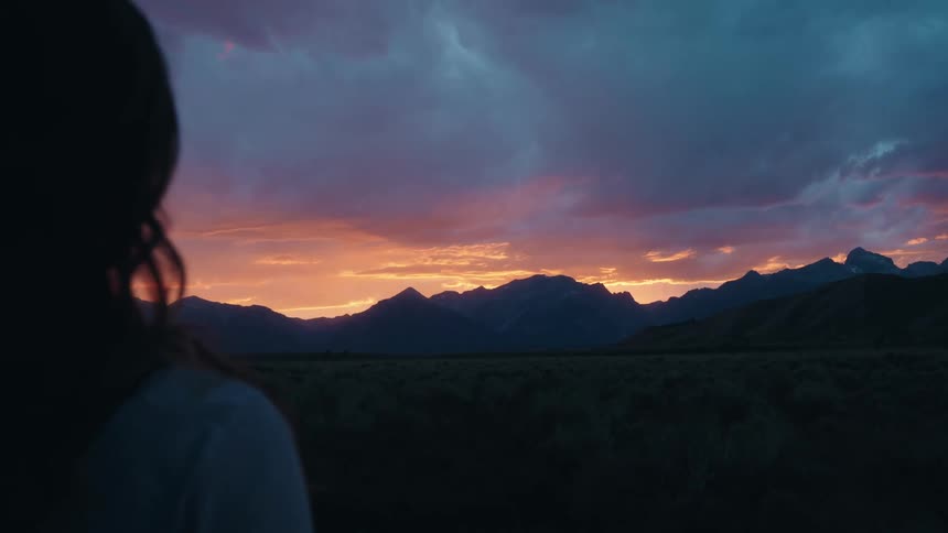 A woman with long hair watches a vibrant sunset over a rugged mountain range silhouetted against the colorful sky