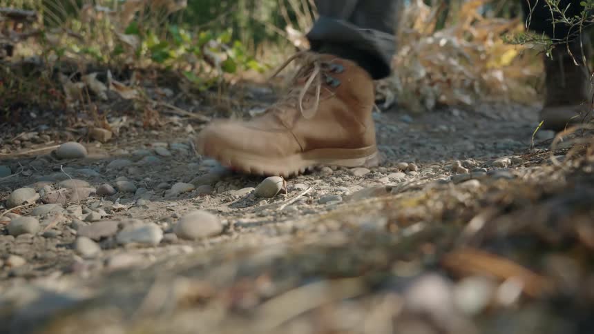 Tan hiking boot steps on rocky ground with dry and green vegetation in natural outdoor setting