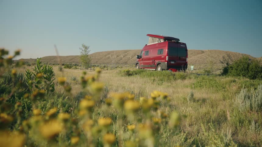 A field of yellow wildflowers captures a red camper van with its poptop roof extended parked on a flat grassy area with rolling arid hills in the background under a bright blue sky