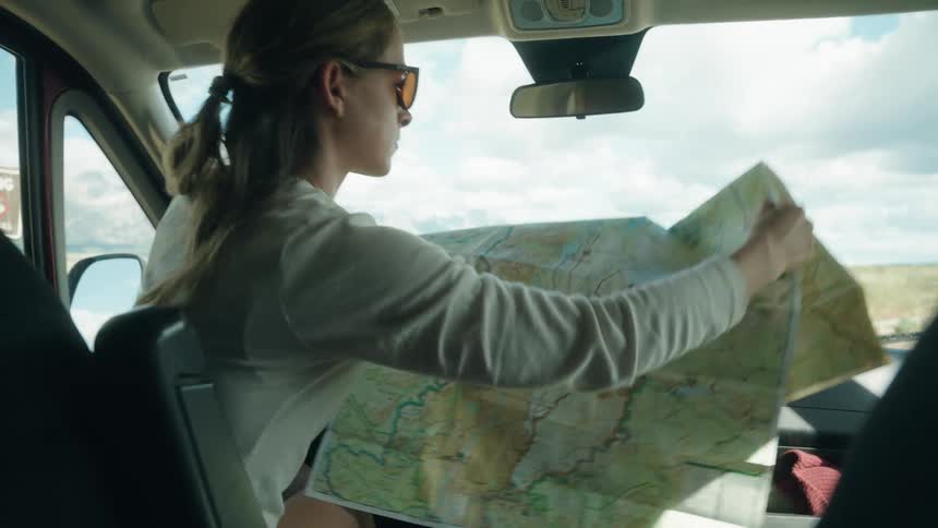 A woman in sunglasses unfolds a large paper map while sitting in the passenger seat of a vehicle