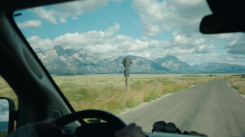 From the drivers perspective a scenic road cuts through a vast plain leading toward a majestic mountain range under a wide clouddappled sky