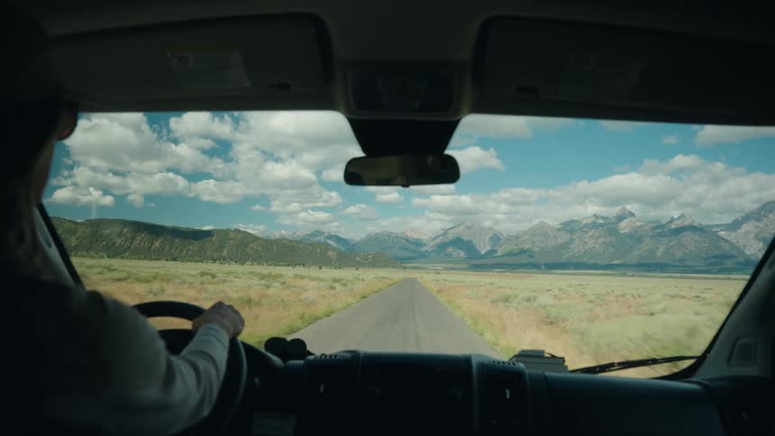 A driver heads down a long straight road toward a majestic mountain range seen from inside a vehicle