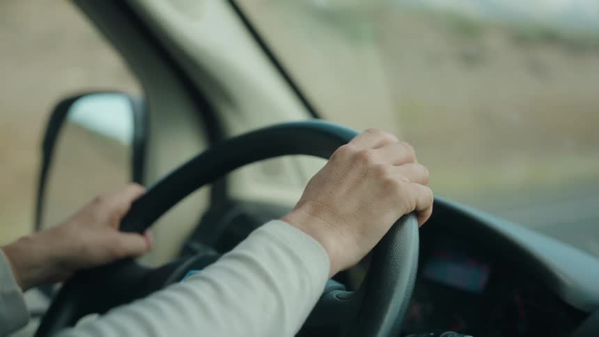 Person driving vehicle with hands on steering wheel and blurred road visible through side window
