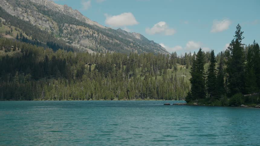 A lake rests at midday surrounded by trees with a mountain range rising in the background
