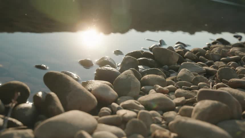 Rounded stones line calm river with golden reflections under low light