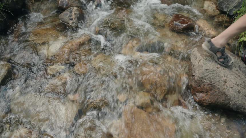 A person walks across rocks above a flowing stream at midday