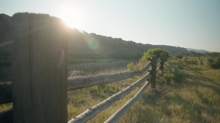Sunlight flares over a rustic wooden fence running along a riverbank backed by rocky cliffs