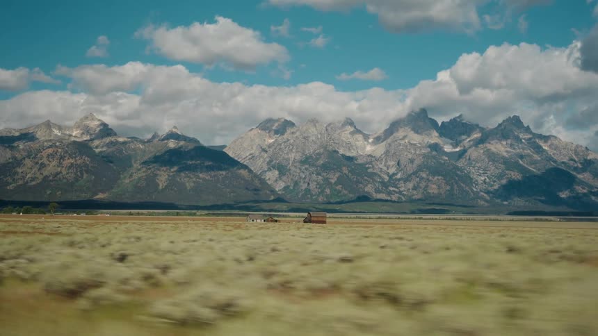 A majestic mountain range rises above a grassy plain with a small barn seen from a moving vehicle