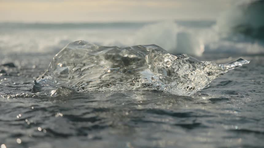 A close view of a transparent ice fragment on dark wet sand with small waves flowing around it and blurred ice shapes visible in the background