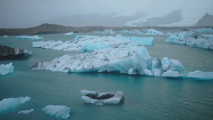 An array of massive sculpted bluetinged icebergs and smaller ice chunks floats in the murky grayblue water of a glacial lagoon under an overcast sky with dark treeless hills forming a rugged backdrop