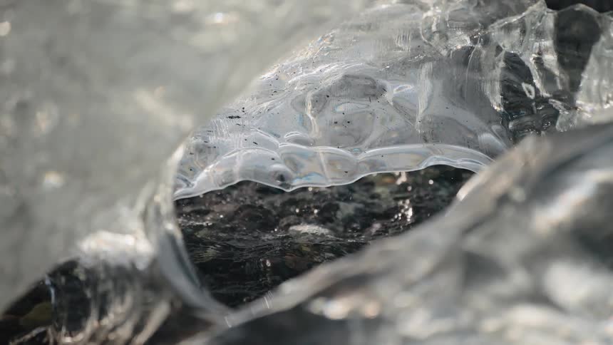 Water washes over dark pebbles visible through an opening in a chunk of clear ice