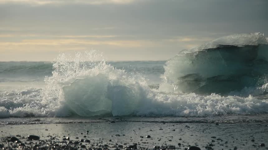 Waves roll in to shore and splash into ice and an iceberg along the coast
