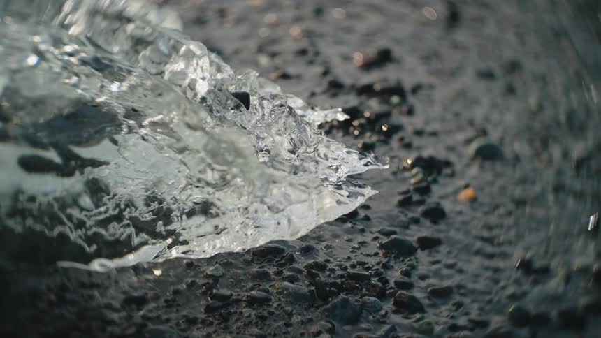 A large chunk of ice rests on the floor of a stream in the wild at midday with water flowing steadily around it